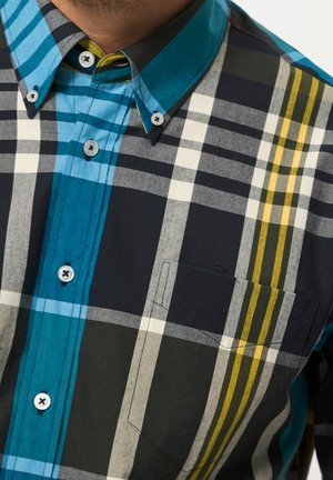 Homme portant une chemise à carreaux multicolore boutonnée avec des rayures bleues, noires, blanches, grises et jaunes et une poche poitrine gauche.