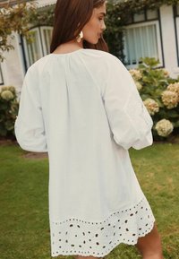 Robe en lin blanc avec des manches bouffantes et un ourlet en forme de vagues comportant des découpes brodées. Texture lisse, coupe décontractée et design simple.