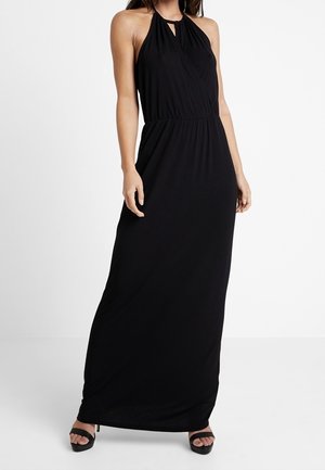 Kobieta ubrana w czarną, bezrękawnikową sukienkę maxi z wiązaniem na szyi, z talią podkreśloną paskiem oraz sandałami na wysokim obcasie i odkrytymi palcami na białym tle.