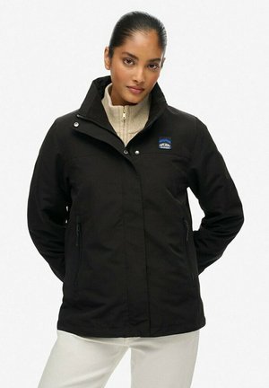Veste noire avec col haut, fermeture zippée et deux poches latérales. Présente un patch logo sur le côté gauche. Texture lisse et mate.
