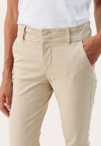 Pantalons kaki en tissu lisse avec un léger brillant, coupe slim, passants de ceinture et poches latérales. Détails de couture à l'avant.