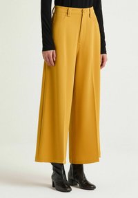 Pantaloni gialli a gamba larga realizzati in materiale liscio, con tasche frontali e chiusura con bottone, abbinati a stivaletti neri con texture.