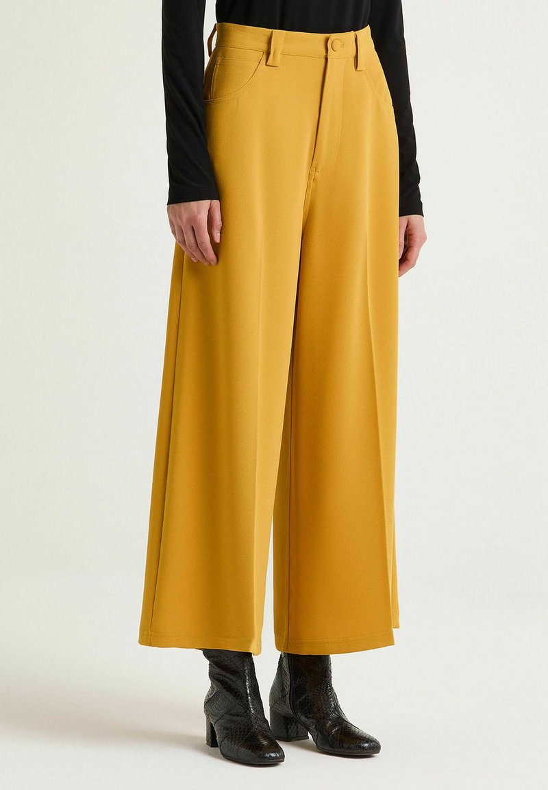 Pantaloni gialli a gamba larga realizzati in materiale liscio, con tasche frontali e chiusura con bottone, abbinati a stivaletti neri con texture.