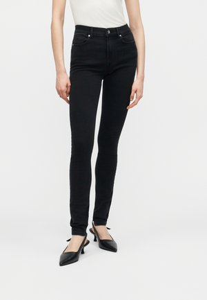 Kvinde iført sort, tætsiddende jeans, hvid ærmeløs top og sorte slingback-hæle med spids tå, stående mod en ensfarvet baggrund.