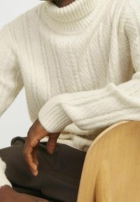 Personne portant un pull à col roulé crème à torsades, assise avec son bras reposant sur une chaise en bois clair.