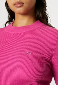 Gros plan d'une femme portant un pull côtelé rose vif avec un petit logo Levi's sur la poitrine.