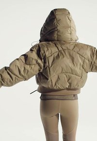 Person trägt eine gepolsterte, braune Kapuzenjacke mit ausgestreckten Armen, kombiniert mit beigefarbenen, eng anliegenden Leggings vor einem einfarbigen hellen Hintergrund.