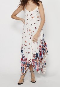 Femme portant une robe blanche sans manches à fleurs avec un ourlet asymétrique et des sandales noires à lanières, se tenant avec une main sur la hanche.
