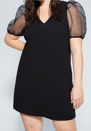 Zwarte jurk met V-hals en doorschijnende pofmouwen met zwarte stippen, gedragen door een persoon met één hand in de zij en golvend donker haar.