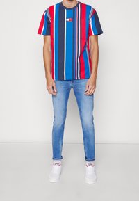 Hombre vistiendo una camiseta de rayas verticales multicolores en rojo, azul y blanco, jeans ajustados de color azul claro y zapatillas blancas con acentos rojos.