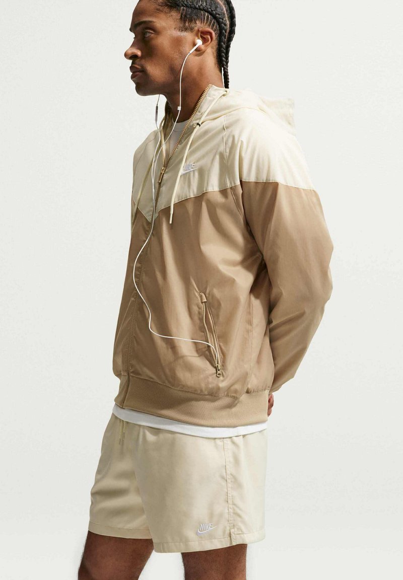 Man met gevlochten haar die een beige en crèmekleurige Nike-jas en korte broek draagt, luistert met witte oortjes naar muziek, staand in profiel.