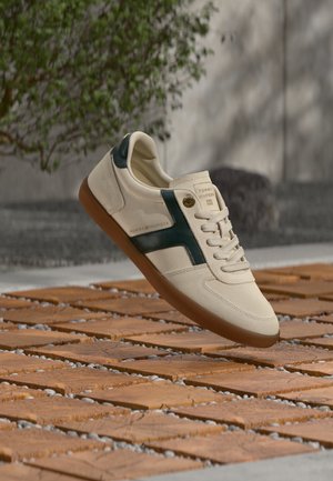 Baskets beiges avec un détail latéral vert foncé et une semelle marron lévitant au-dessus d’un sol en carrelage en bois à l’extérieur avec de la verdure en arrière-plan.