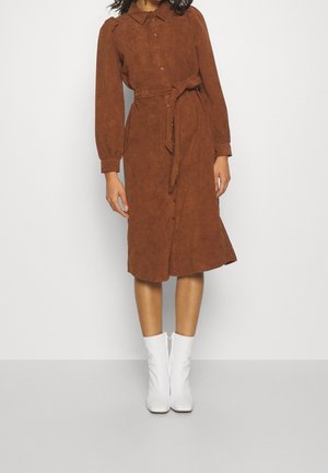 Brązowa sukienka z sztruksu o średniej długości, z długimi rękawami, z przodu na guziki, z wiązanym paskiem w talii, noszona z białymi botkami za kostkę.