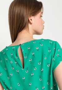 Blusa verde com padrão floral, decote redondo, mangas curtas e pequeno fecho de botão nas costas. Tecido suave e texturizado.