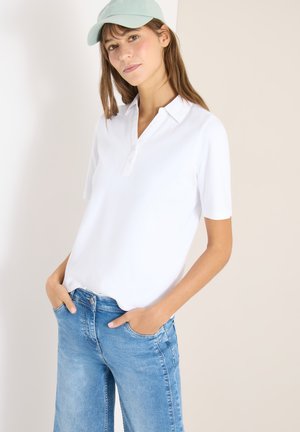 Vrouw draagt een lichtblauwe pet, een wit kort mouws overhemd met kraag en een blauwe spijkerbroek, met handen in de zakken, staand tegen een effen achtergrond.