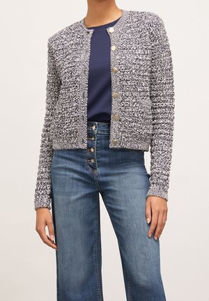 Femme portant un cardigan texturé noir et blanc boutonné sur une chemise bleu marine et un jean bleu taille haute avec plusieurs boutons.