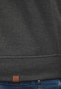 Mørkegrå sweatshirtstof med ribbet kant og et lille brunt lædermærke med et globusdesign. Teksturen ser blød og glat ud.
