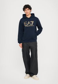 Mørk blå hettegenser med gulltrykt logo, med kengurulomme, kombinert med vide svarte cargo-bukser og beige sneakers.