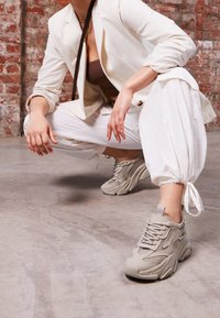 Osoba w squacie, ubrana w białe spodnie, beżowe masywne sneakersy, białą marynarkę i brązowy top, na tle ceglanej ściany i betonowej podłogi.