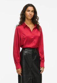 Woman with long curly hair wearing a red satin button-up blouse tucked into a high-waisted black leather skirt against a white background.