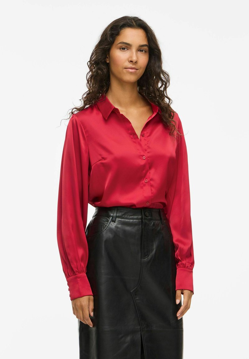 Woman with long curly hair wearing a red satin button-up blouse tucked into a high-waisted black leather skirt against a white background.