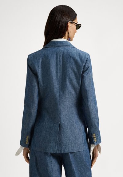 Tmavomodrý rifľový blejzer s prispôsobeným strihom, výstrihmi s výrezmi a gombíkovými manžetami. Viditeľný detail švov a rozpark na zadnej strane.