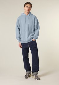 Felpa oversize azzurra chiara con cordini e tasca frontale, abbinata a jeans in denim scuro e sneakers grigie camo.