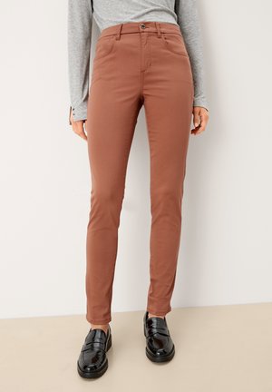 Woman wearing slim-fit rust-colored pants with front pockets and black loafers, paired with a light gray long-sleeve top.