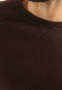 Sudadera de algodón marrón con cuello redondo, con detalles de costura y un escote acanalado. Presenta una textura suave y una vista de cerca de la elasticidad de la tela.