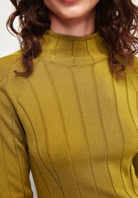 Olivenfarbener, gerippter Strickpullover mit Stehkragen, der über vertikale Streifen, eine strukturierte Oberfläche und einen hohen Kragen verfügt.