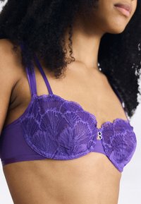 Mulher vestindo um sutiã de renda roxa com alças duplas, mostrada dos ombros até a metade do torso contra um fundo simples.