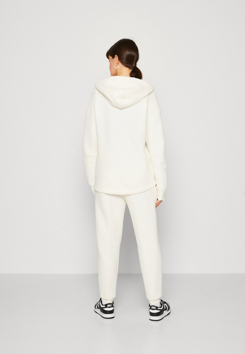 Sudadera blanca con una gran capucha, combinada con pantalones a juego. Textura suave, corte relajado, con hombros caídos y puños acanalados. Zapatillas negras.