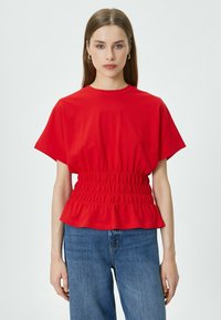 T-shirt rosso realizzato in tessuto morbido, caratterizzato da una silhouette ampia, vita arricciata con orlo a volant e maniche corte. Abbinato a jeans blu.