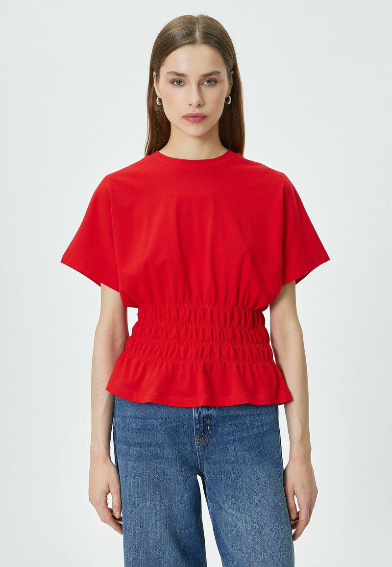 T-shirt rosso realizzato in tessuto morbido, caratterizzato da una silhouette ampia, vita arricciata con orlo a volant e maniche corte. Abbinato a jeans blu.