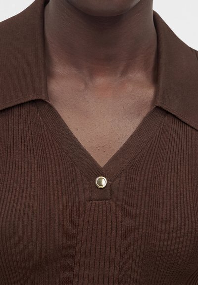 Κοντινό πλάνο ενός ατόμου που φοράει καφέ ριμπ πλεκτό τοπ με V-λαιμόκοψη και ένα μικρό στρογγυλό χρυσό κουμπί κάτω από το γιακά.