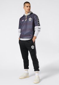 Hombre vistiendo una camiseta deportiva a rayas negras y moradas sobre una sudadera gris, pantalones de chándal negros con logo, calcetines blancos y zapatillas blancas, de pie con las manos en los bolsillos.