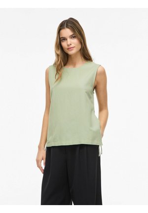 Młoda kobieta o długich włosach, ubrana w bezrękawnik jasnozielony top i luźne czarne spodnie na jednolitym białym tle.