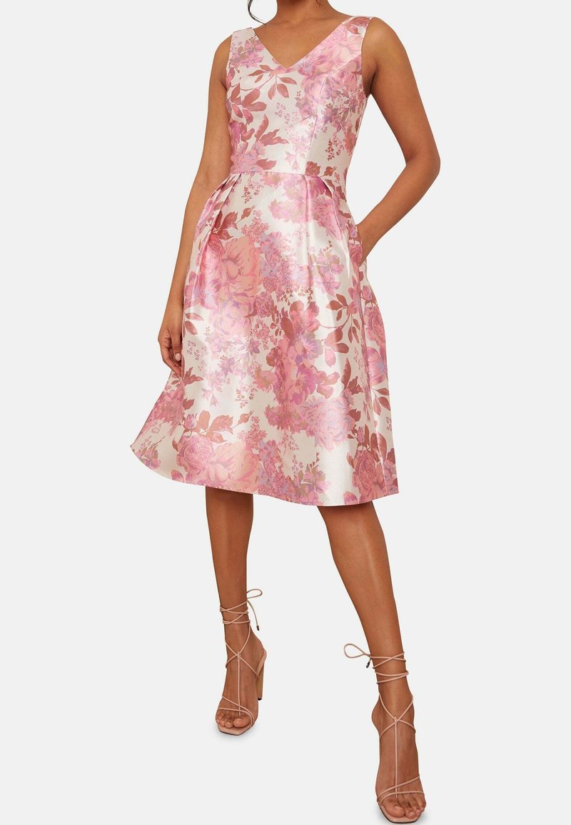 Femme portant une robe rose à fleurs, sans manches, longueur genou, avec un col en V et des poches, assortie à des sandales à talons hauts à brides.