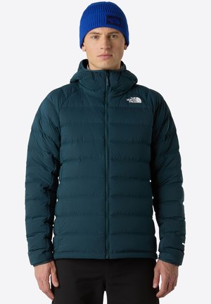 Dark teal puffer jacket with a zip front, hood, and ribbed cuffs. Features white logo on the upper left. Accompanied by a blue knit beanie.
