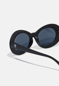 Gafas de sol negras con lentes redondos y acabado mate. La montura tiene una forma audaz, con lados gruesos y un logotipo de acento en la patilla.