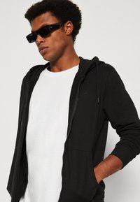 Schwarze Zip-Hoodie mit weicher Textur, ausgestattet mit einer Kängurutasche und Kordelzügen. Getragen über einem weißen T-Shirt, ergänzt durch dunkle Sonnenbrillen.