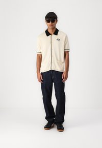 Cremfarbener Cardigan mit kurzen Ärmeln und schwarzem Kragen, verziert mit einem Logo, kombiniert mit dunklen Jeans und schwarzen Sneakers. Einfaches und lässiges Design.