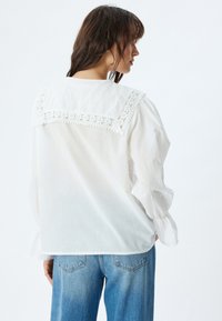 Witte blouse met een met kant afgewerkt schouderstuk, lange, ballonachtige mouwen en een zachte, lichte textuur. Gecombineerd met lichtblauwe high-waisted jeans.