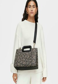 Mujer vestida con un suéter blanco de gran tamaño y pantalones, llevando un bolso rectangular estampado con un asa negra y una correa ancha para el hombro.