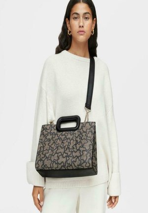 Mujer vestida con un suéter blanco de gran tamaño y pantalones, llevando un bolso rectangular estampado con un asa negra y una correa ancha para el hombro.