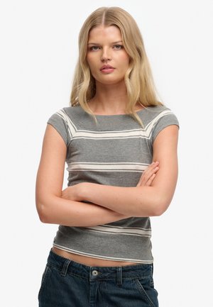 Femme blonde aux cheveux raides portant un haut à manches courtes gris rayé et un jean bleu, les bras croisés, expression neutre.