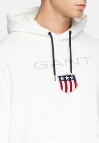 Weißer Hoodie mit blauen Kordeln, der ein auffälliges gesticktes Wappen mit rot, weiß und blauen Streifen zeigt. Das Logo lautet "GANT."