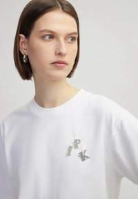 Witte katoenen t-shirt met korte mouwen, met glanzende juweelachtige letters "I," "P," en "K" in een verspreide opstelling op de voorkant.