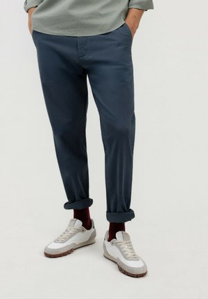 Hombre que lleva pantalones azul marino remangados, calcetines burdeos, zapatillas blancas y una camisa de manga larga verde claro con las manos en los bolsillos.