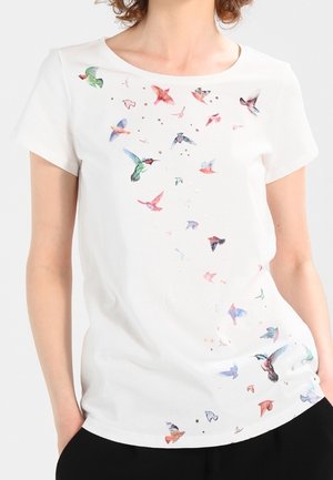 Chemise blanche à manches courtes avec de petits oiseaux colorés dispersés en diagonale sur le devant, portée par une personne les mains dans les poches.